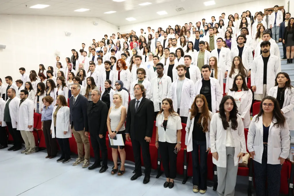 Amfinin basamaklarında ayakta duran beyaz önlüklü öğrenciler ve ön sırada öğretim üyeleri