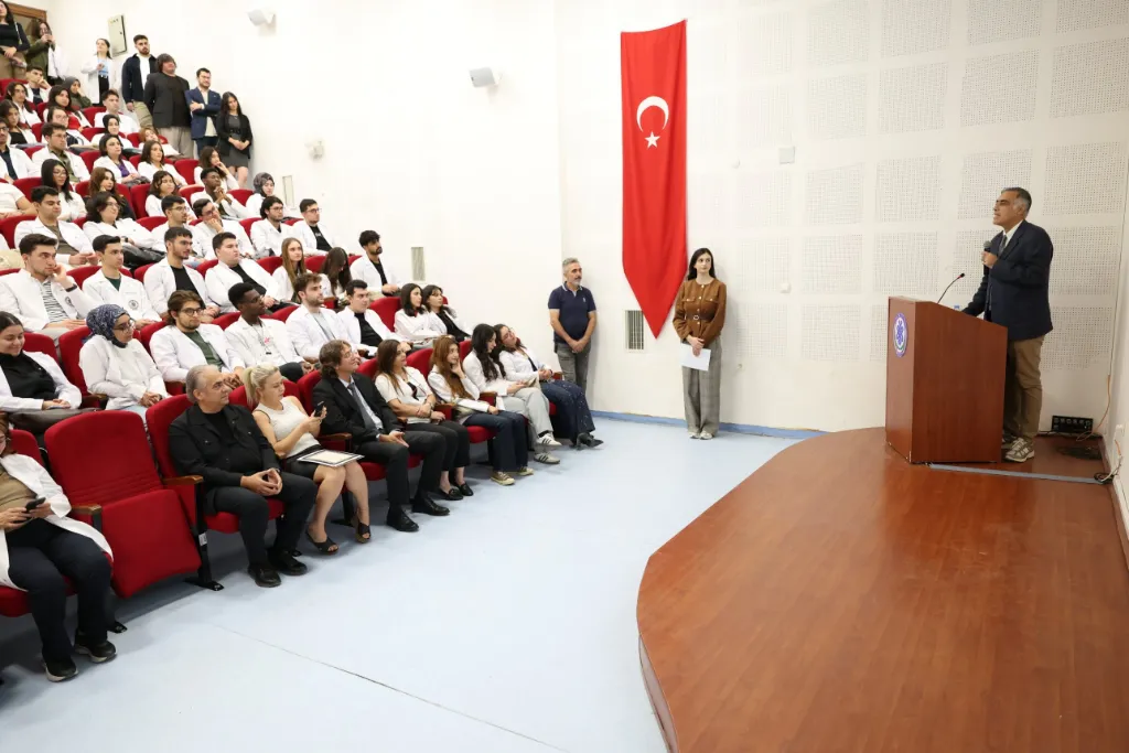 Amfide oturan öğrenciler ve öğretim üyelerine kürsüden seslenen Dekan; duvarda Türk bayrağı